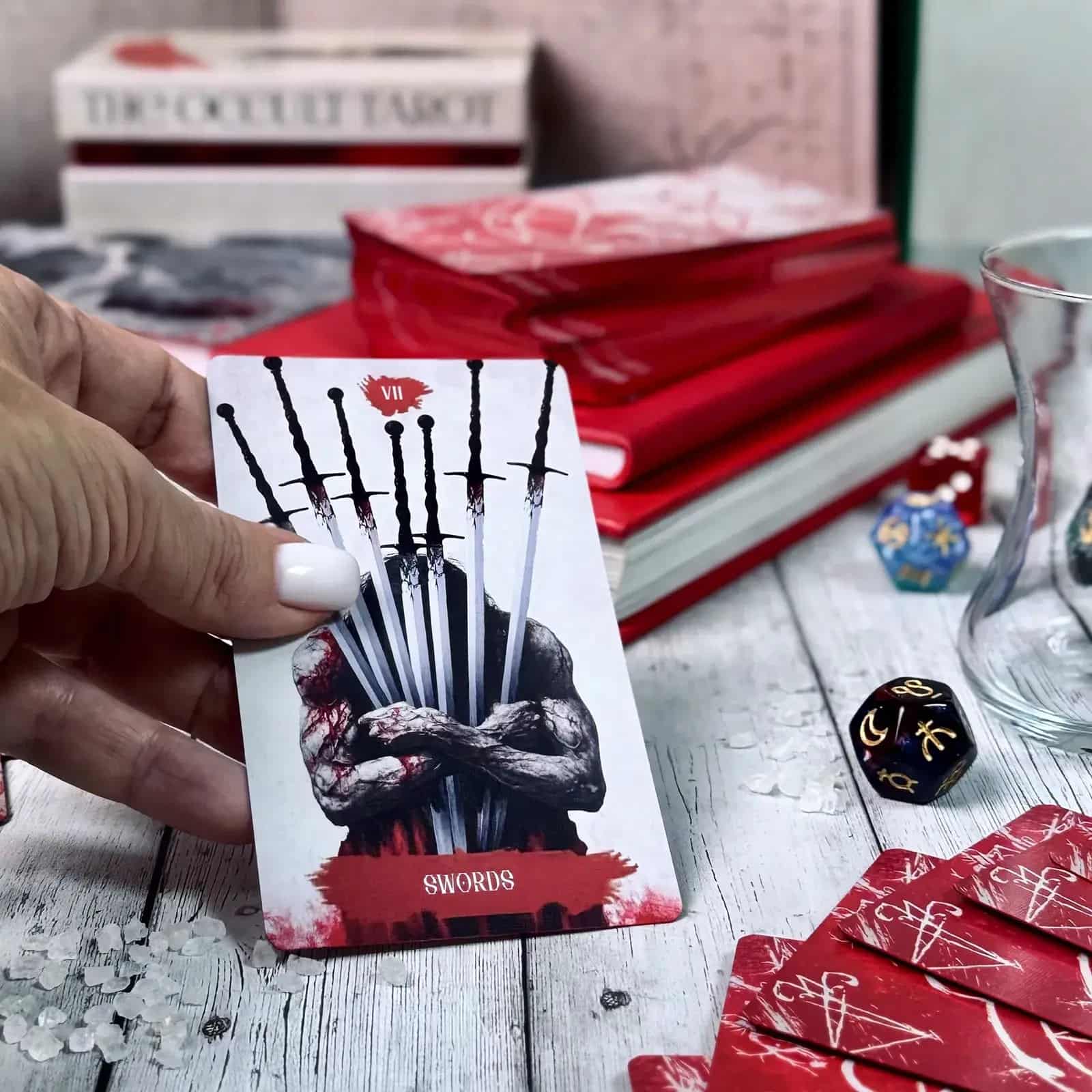 Arcana Obscura Seven of Swords tarot card held in hand with red deck dice and reading setup