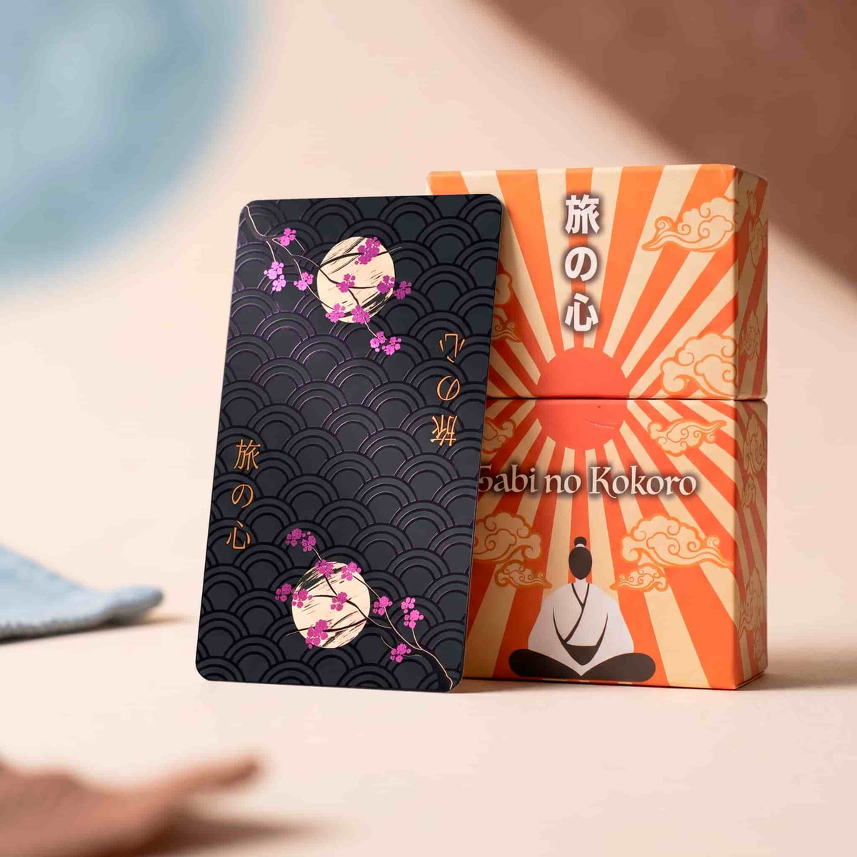Tabi no Kokoro Tarot black card back with pink cherry blossom foil beside orange rising sun collector box