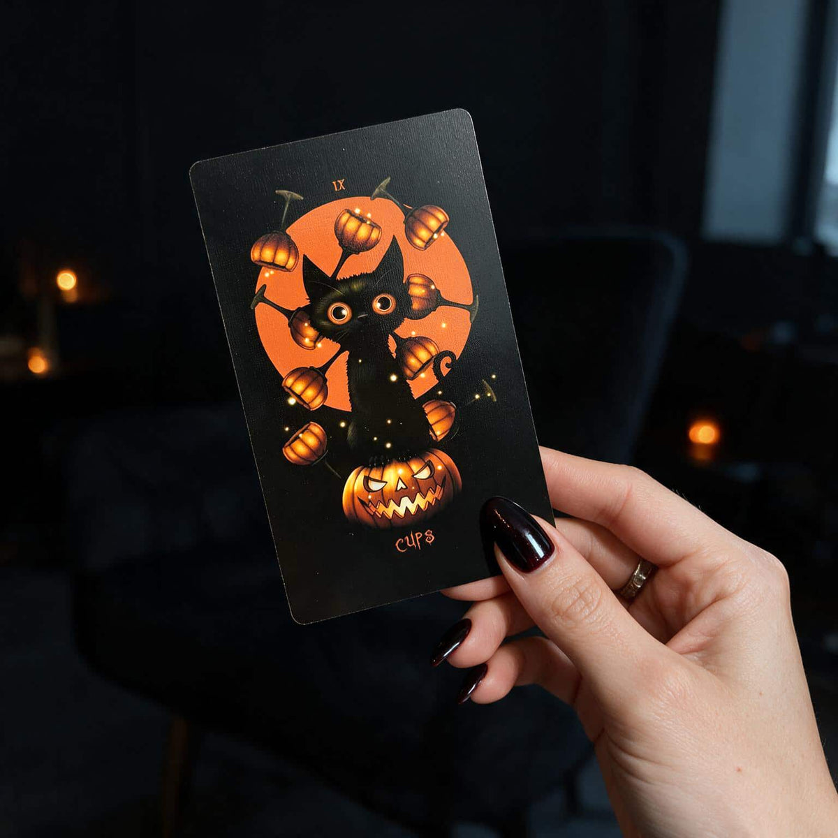 Hand with black nails holding Halloween Tarot Nine of Cups card, black cat with pumpkin cups