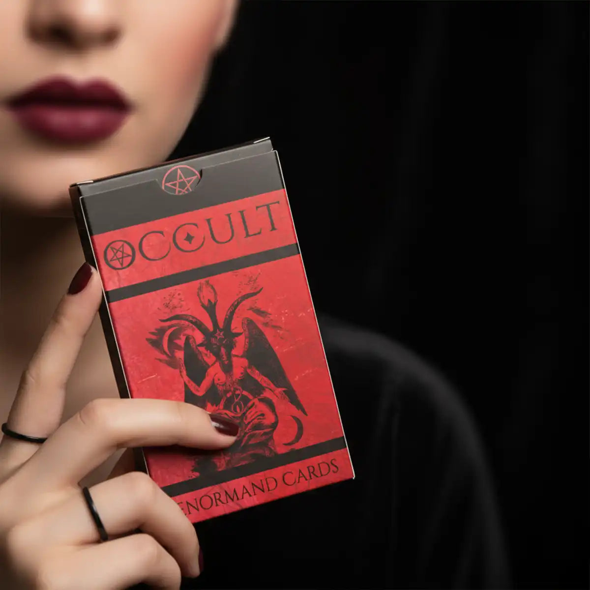 Occult Lenormand deck box with red and black design held by woman with dark aesthetic makeup on black background