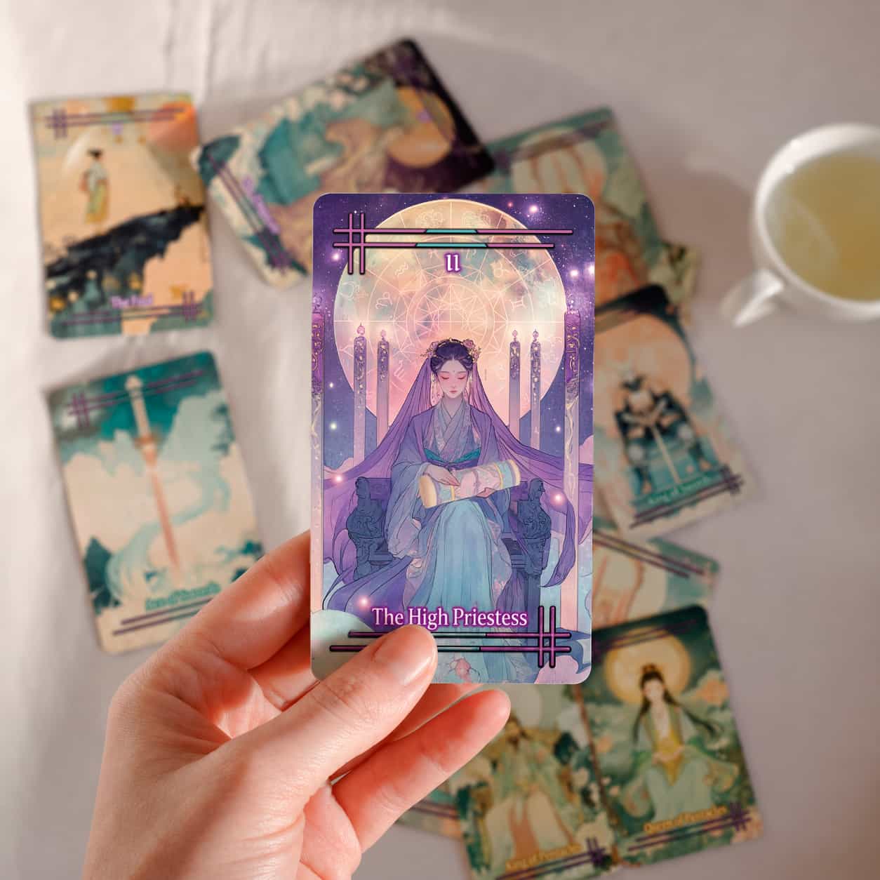 Hand holding a tarot card titled 'The High Priestess' with other tarot cards and a cup of tea on a white surface.