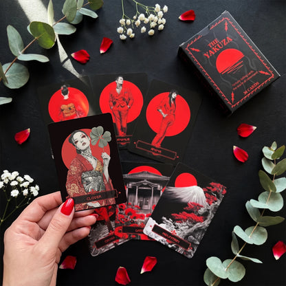 Yakuza Lenormand cards flat lay with red and black design, hand holding clover card, surrounded by petals and greenery